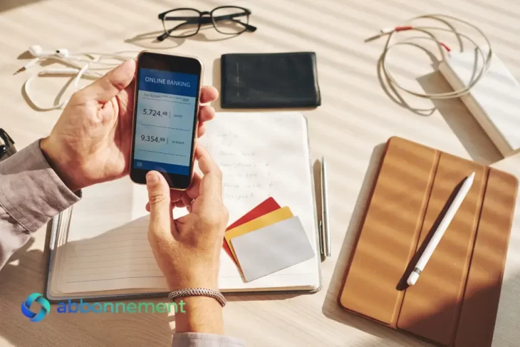 Persona che controlla il Conto Corrente Online dallo smartphone, con carte e taccuino su una scrivania.