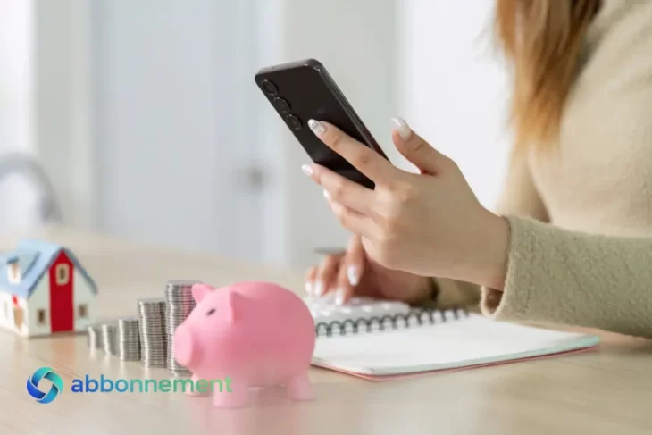 Persona che controlla le spese dallo smartphone con salvadanaio e monete sul tavolo, esempio di Gestire le Spese.