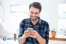 Uomo sorridente seduto in una stanza luminosa mentre guarda il suo smartphone per controllare l’app di budgeting e organizzare le spese quotidiane.