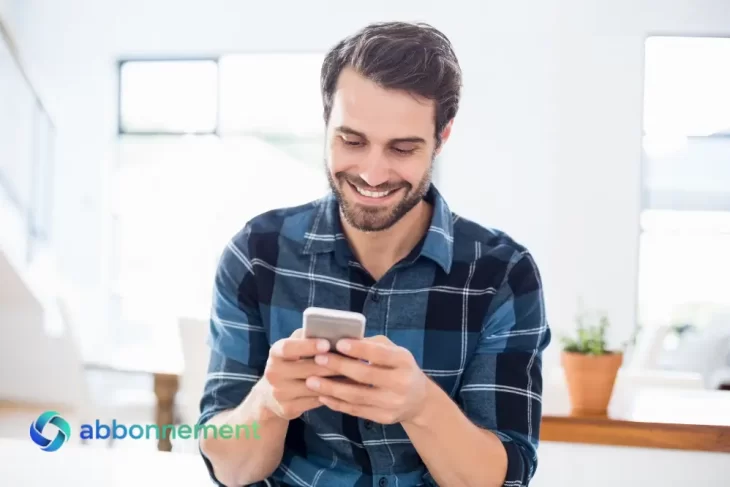 Uomo sorridente seduto in una stanza luminosa mentre guarda il suo smartphone per controllare l’app di budgeting e organizzare le spese quotidiane.