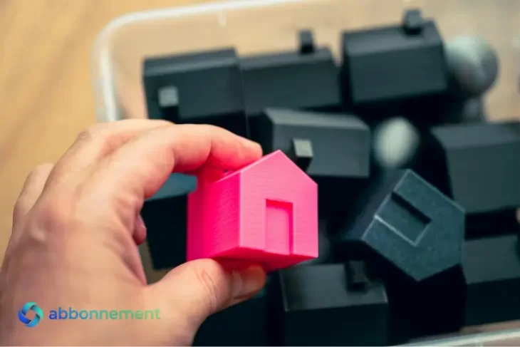Mano eligiendo una casa rosa entre varias, representando la decisión de comprar o alquilar vivienda.